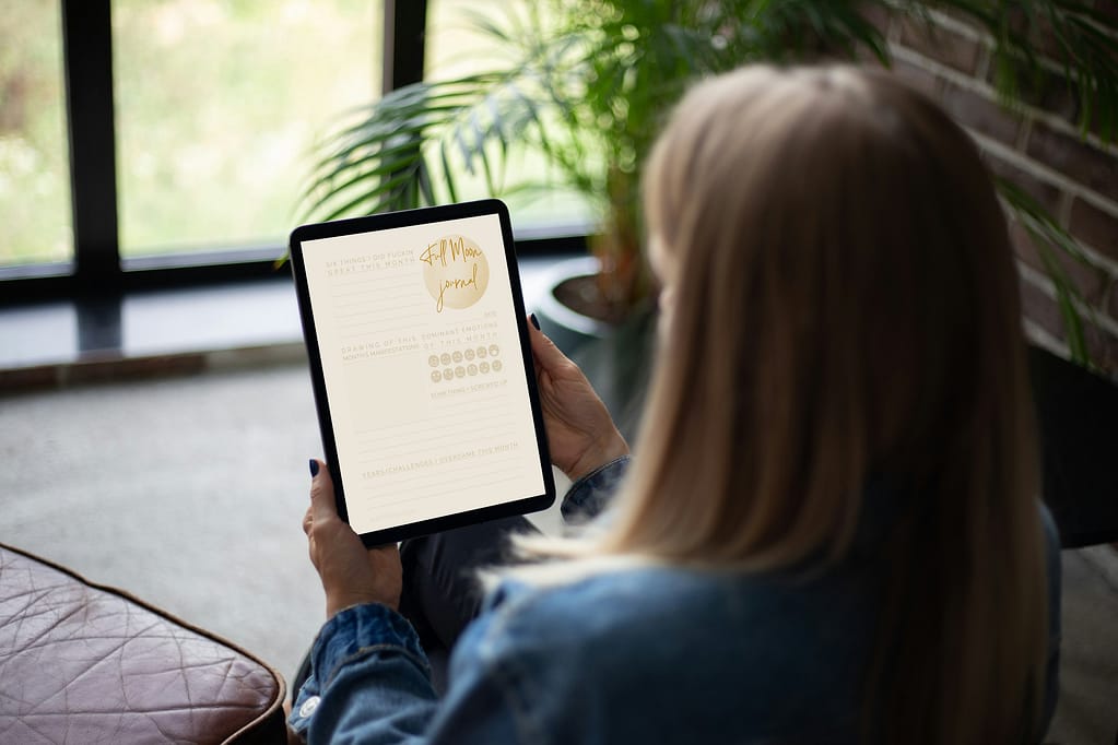 A person sitting by a window, holding a tablet displaying the ‘Full Moon Journal,’ perfect for reflecting on completions and letting go.