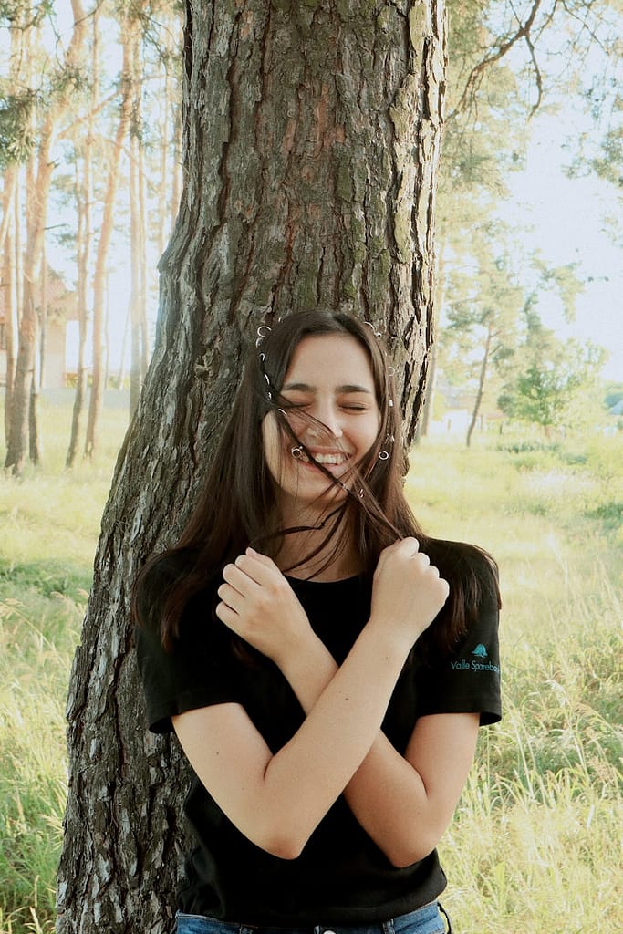 Smiling woman standing against a tree with arms crossed, embodying self-connection and joy — a moment of healing through tarot and personal clarity.