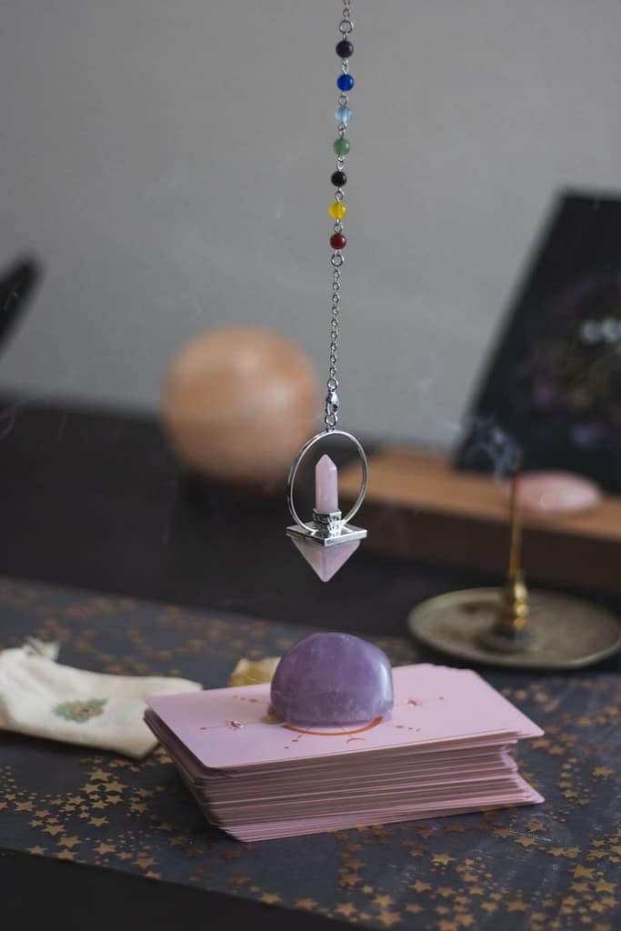A pendulum with colored beads hangs over stacked tarot cards, creating a mystical ambiance.