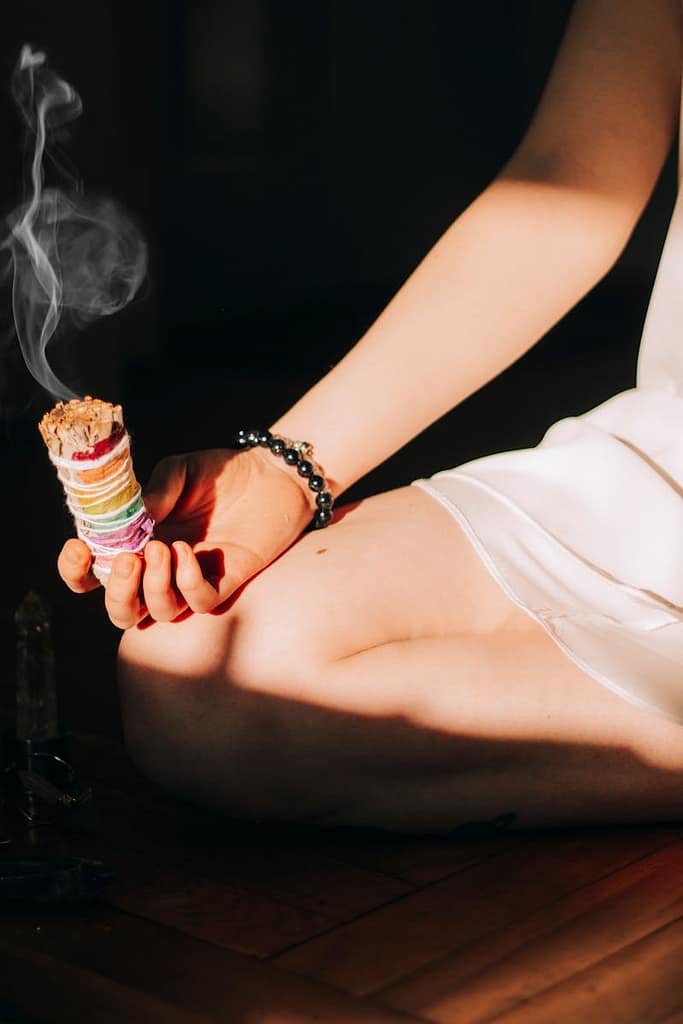 Woman meditates with a colorful smudge stick in hand, emitting smoke indoors.