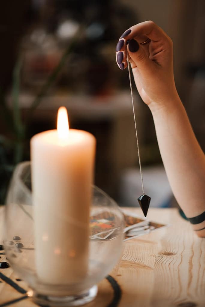 A hand holds a pendulum over tarot cards by candlelight, evoking a mystical ambiance.