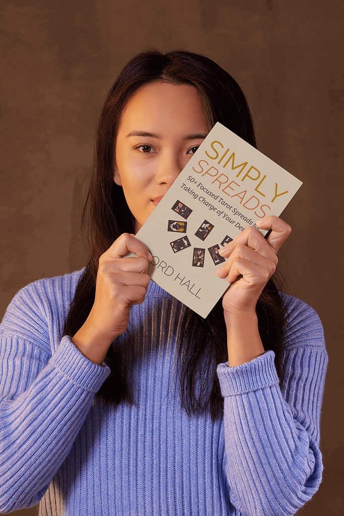 A woman in a blue sweater holding up the book “Simply Spreads” by Gord Hall in front of her face, showcasing its cover. The book features tarot cards on the cover and highlights its focus on tarot spreads for self-empowerment.