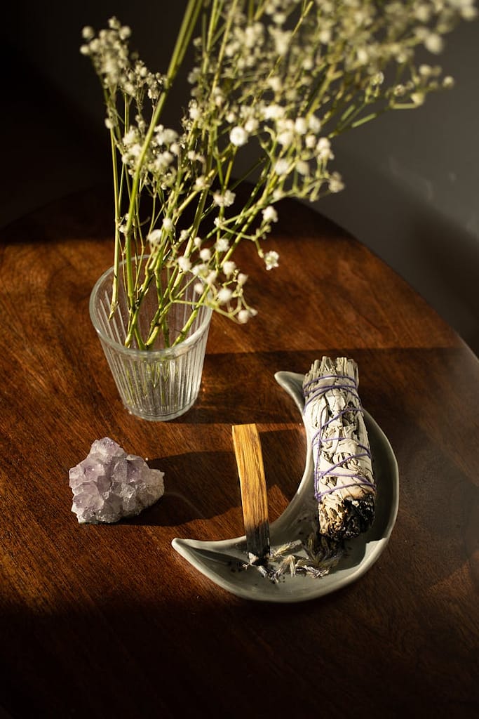 Still life of white sage bundle, palo santo, amethyst, flowers, and incense on a wooden table.