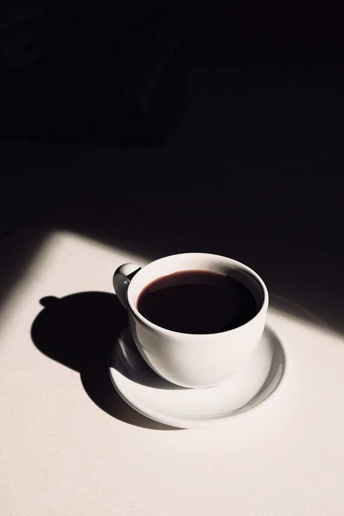 A single white cup of black coffee casting a shadow on a light surface, symbolising a moment of pause, reflection, and intentional emotional refilling.
