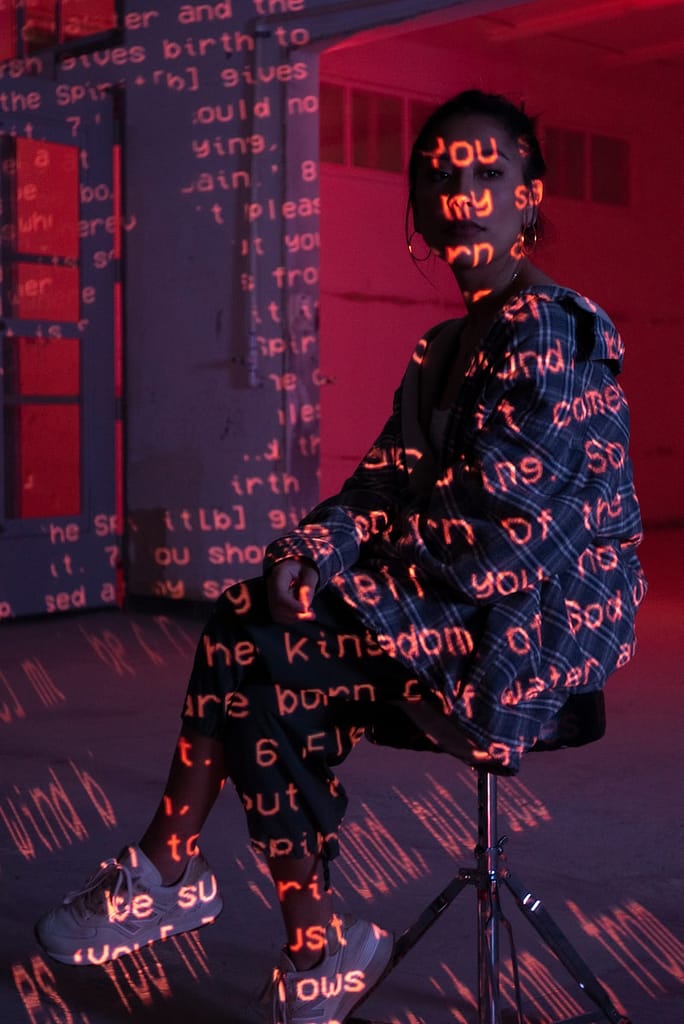 Person sitting in a dark room with red text projected across their body, symbolising carrying lies as truth and the hidden stories we absorb.