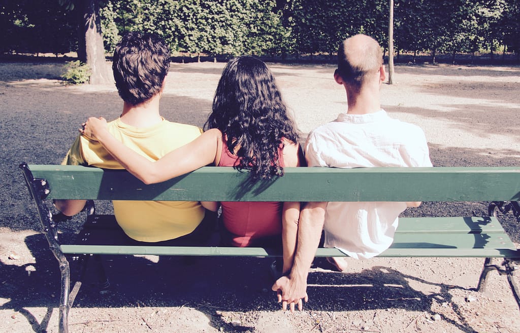 Find The Best Tarot Spread For Love, Sex, And Relationships 8 Three people sitting on a park bench. The person in the middle has their arms around both individuals on either side, symbolizing a polyamorous relationship. One person on the end is holding hands secretly with the person in the middle, highlighting the complexity of relationships.
