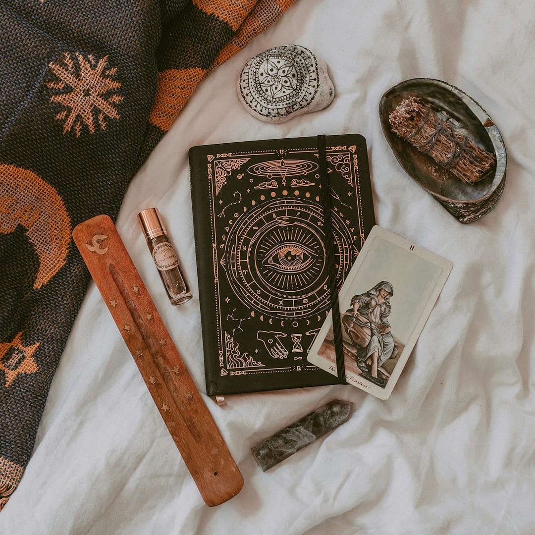 Flat lay of spiritual tools including a tarot card, black journal with mystical design, incense holder, sage bundle, and crystals, representing tarot, journalling and meditation in Manchester.