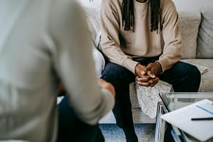 Unrecognizable African American male patient with clasped hands sitting on sofa near blurred anonymous therapist during visit in psychotherapy office