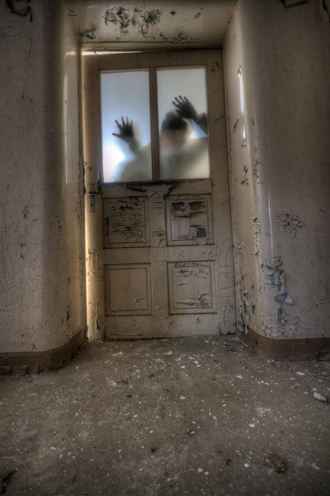 How To Spot A Fake Tarot Reader Disturbing image symbolising how to spot a fake tarot reader, showing a shadowy figure behind a broken door in an eerie, abandoned hallway.