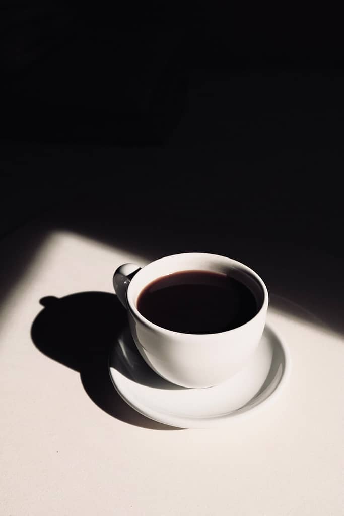 A single white cup of black coffee casting a shadow on a light surface, symbolising a moment of pause, reflection, and intentional emotional refilling.
