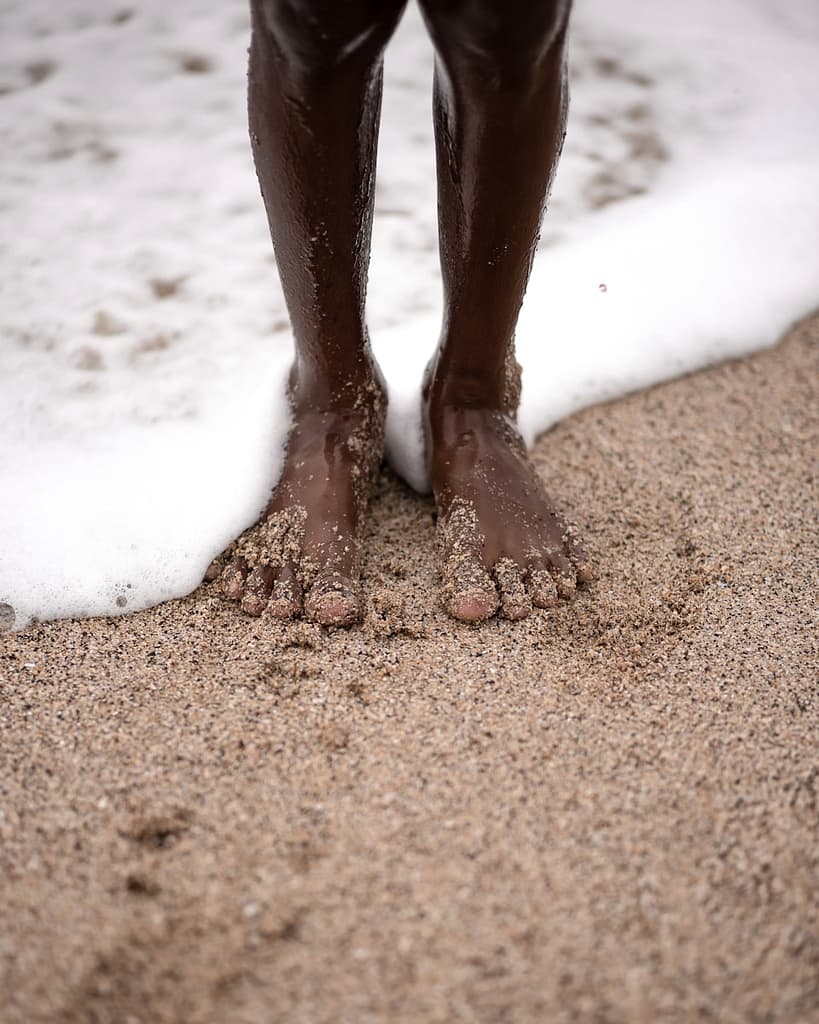 Close-up of bare feet on wet sand at the edge of the sea, symbolising grounding practices and the earthy stability of the suit of Pentacles.