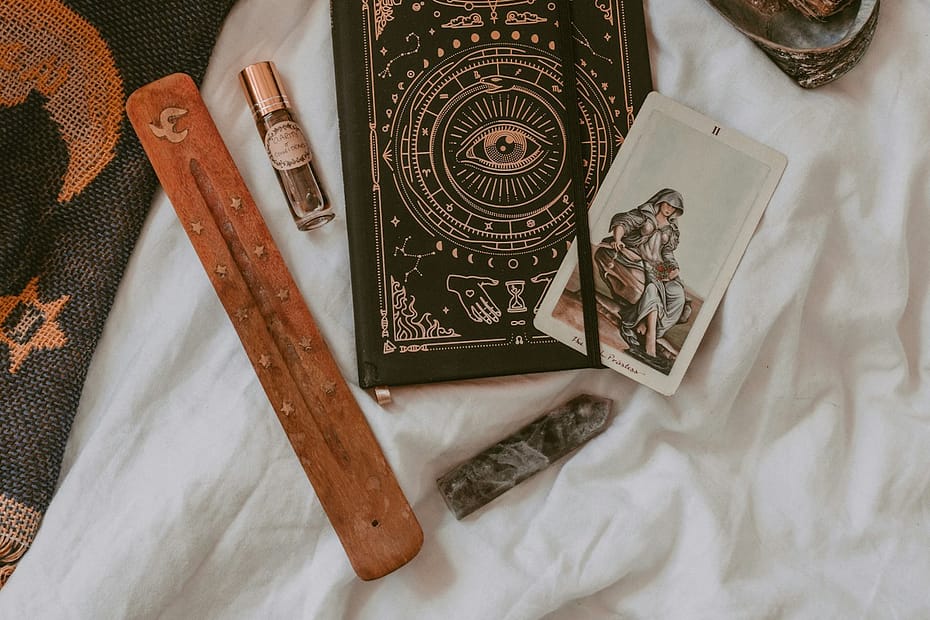 Flat lay of spiritual tools including a tarot card, black journal with mystical design, incense holder, sage bundle, and crystals, representing tarot, journalling and meditation in Manchester.